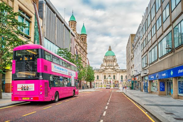 Bus stands at a bus stop with The City Hall building in the background in downtown Belfast Northern Ireland on a cloudy day.. Public Transport Statistics Northern Ireland 2022-23